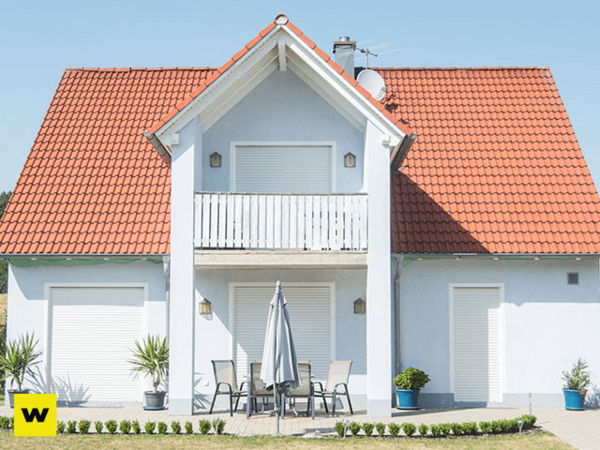 Modernes Einfamilienhaus mit rotem Dach, Balkon und geschlossener Terrasse im Sonnenschein.